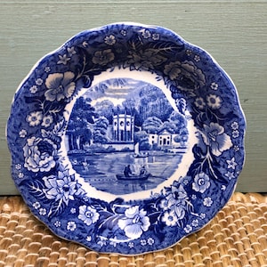 May include: Blue and white decorative plate with a floral border. The center features a scenic landscape with a lake, buildings, and people in a boat. The plate has a scalloped edge.