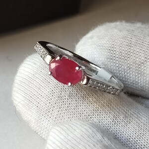 May include: A silver ring featuring a vivid red oval gemstone. The ring's band is embellished with small, clear accent stones. The ring is presented on a white surface.