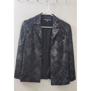 May include: A black blazer featuring a dark grey patchwork pattern with intricate floral and geometric designs. The garment has a notched lapel, long sleeves, and a solid black interior lining. The label inside the collar reads Christine Foley.
