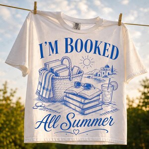 Op de afbeelding: Een wit t-shirt met een blauwe grafische print met de tekst I'M BOOKED All Summer. Het ontwerp bevat een gevlochten mand vol boeken, een stapel boeken met een zonnebril erbovenop, een glas met een rietje en een kustlandschap met gebouwen.