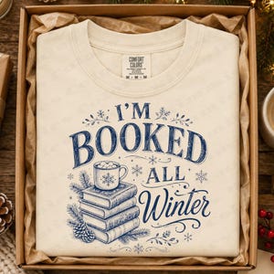 May include: A cream-colored t-shirt featuring a dark blue graphic print that reads I'M BOOKED ALL WINTER. The design includes a stack of three books topped with a mug of hot cocoa, surrounded by pine branches, snowflakes, and decorative flourishes.