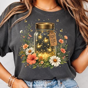 May include: Dark gray t-shirt featuring a mason jar filled with glowing fireflies. The jar is surrounded by colorful flowers and greenery. A tag on the jar reads "Summer Nights & Firefly Lights."