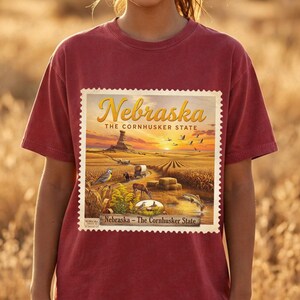 Op de afbeelding: Een bordeauxrood t-shirt met een Nebraska staatsafbeelding. De afbeelding, die lijkt op een vintage postzegel, toont een zonsondergang boven een landelijk landschap met de tekst "Nebraska - The Cornhusker State". Het ontwerp bevat velden, dieren in het wild en een butte.
