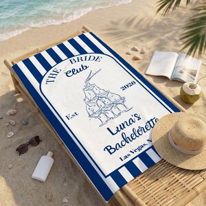 May include: A white beach towel with blue stripes and a central design. The design includes the text "THE BRIDE Club" and "Luna's Bachelorette" with a champagne glass tower illustration. Other items include sunglasses, a hat, and a book.