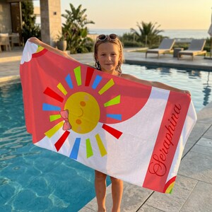 May include: A beach towel with a colorful sun design and the name "Seraphina" in red script. The towel features a red border and a white background. The sun has a yellow face, a pink bow, and colorful rays.