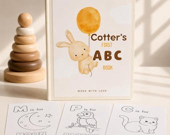 Peut inclure: Un livre relié intitulé Cotter's First ABC Book avec une illustration à l'aquarelle d'un lapin tenant un ballon jaune, accompagné de trois cartes d'activités blanches avec des dessins au trait noir d'une lune, d'un perroquet et d'un chat.