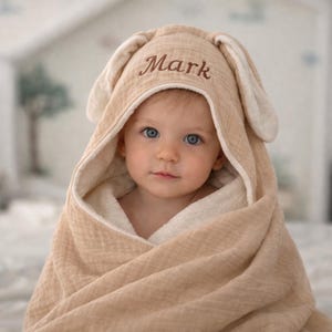 May include: A baby wrapped in a tan hooded towel with bunny ears. The towel has a white trim and the name "Mark" embroidered on the hood in brown. The baby has blue eyes and is looking at the camera.