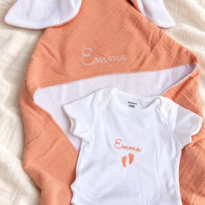 Peut inclure: Une serviette &agrave; capuche couleur p&ecirc;che avec des oreilles de lapin blanches et le pr&eacute;nom Emma brod&eacute; en blanc, accompagn&eacute;e d'un body blanc &agrave; manches courtes avec le pr&eacute;nom Emma et deux petites empreintes de pieds couleur p&ecirc;che brod&eacute;es.