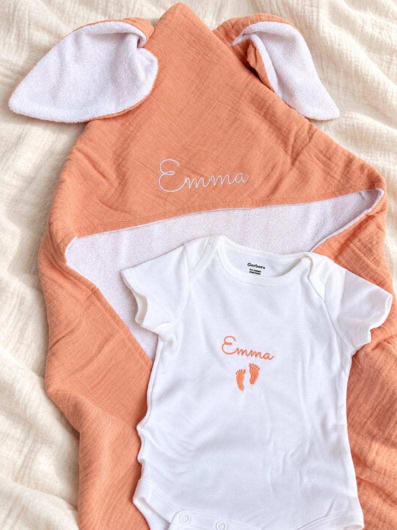 Peut inclure: Une serviette &agrave; capuche couleur p&ecirc;che avec des oreilles de lapin blanches et le pr&eacute;nom Emma brod&eacute; en blanc, accompagn&eacute;e d'un body blanc &agrave; manches courtes avec le pr&eacute;nom Emma et deux petites empreintes de pieds couleur p&ecirc;che brod&eacute;es.