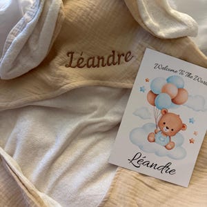 Peut inclure: Une serviette &agrave; capuche beige avec des oreilles de lapin, brod&eacute;e du nom L&eacute;andre en fil marron. &Agrave; c&ocirc;t&eacute; se trouve une carte blanche illustr&eacute;e d'un ours en peluche tenant des ballons bleus et p&ecirc;che, avec le texte Welcome To The World et L&eacute;andre.