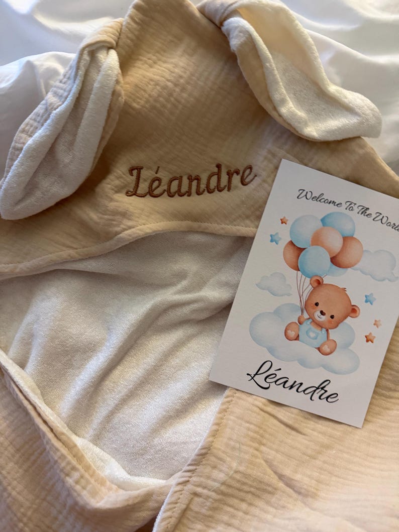 Peut inclure: Une serviette &agrave; capuche beige avec des oreilles de lapin, brod&eacute;e du nom L&eacute;andre en fil marron. &Agrave; c&ocirc;t&eacute; se trouve une carte blanche illustr&eacute;e d'un ours en peluche tenant des ballons bleus et p&ecirc;che, avec le texte Welcome To The World et L&eacute;andre.