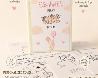 Peut inclure: Un livre ABC personnalisé de 13 x 18 cm pour bébés avec une couverture claire illustrée d'un lapin tenant un ballon rose, des nuages et des papillons. Le texte sur la couverture indique Elisabeth's FIRST ABC BOOK. Plusieurs cartes blanches avec des dessins au trait noir d'objets comme un biberon, un éléphant et un cerf-volant sont présentées au premier plan.