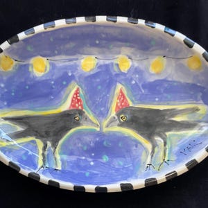 May include: An oval ceramic dish with a black and white striped rim. The interior features a blue background with two black birds wearing red and white polka-dot party hats, facing each other under a string of yellow lights. The artist signature is visible.