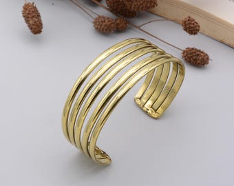 May include: A gold-colored cuff bracelet with six parallel bands. The bracelet has a hammered texture and an open design. The jewelry is displayed on a light gray surface, with dried floral elements in the background.