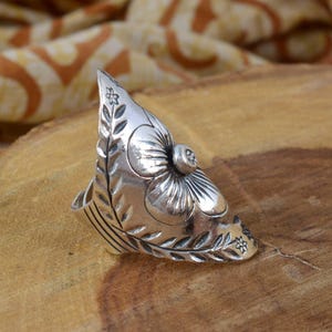 May include: A silver-colored ring with a floral design. The ring features a large flower with detailed petals and a smaller flower on the side. The ring is set on a wooden surface.