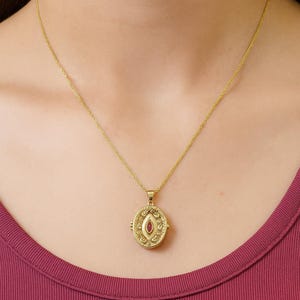 May include: Gold-coloured pendant necklace with a small, oval-shaped locket. The locket features an ornate design and a red gemstone in the centre. The necklace is worn against a maroon top.