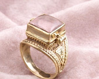 May include: A gold-colored ring with a large, rectangular, light pink gemstone. The ring has an ornate design on the band and around the gemstone setting. The ring is set against a soft pink background.