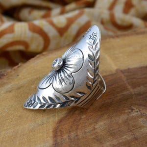 May include: A silver-toned ring with a floral design. The ring features a large flower on the front and leaf patterns along the sides. The ring is set on a wooden surface.
