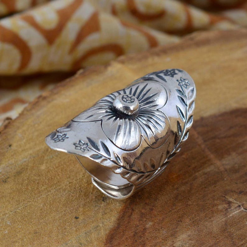 May include: A silver-toned ring with a floral design. The ring features a large flower on top with detailed petals and a central stud. The band is adorned with smaller floral and leaf patterns. The ring is set against a wooden surface.