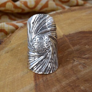 Könnte beinhalten: Ein silberfarbener Ring mit einem detaillierten Spiraldesign. Der Ring hat ein zentrales Blumenmotiv und strukturierte, gebogene Linien, die nach außen strahlen. Der Ring liegt auf einer Holzoberfläche.