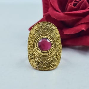 Puede incluir: Un anillo dorado con forma ovalada y intrincados diseños de volutas. El anillo presenta una piedra grande, redonda, de color rubí en el centro. El fondo incluye una rosa roja, sugiriendo un tema romántico.