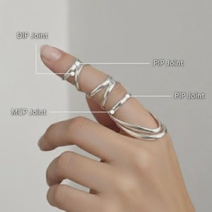 Op de afbeelding: Een zilverkleurige vingerring met een uniek ontwerp. De ring wikkelt zich om de vinger en bedekt de DIP-, PIP- en MCP-gewrichten. De ring heeft een reeks gebogen en rechte lijnen, waardoor een ingewikkelde en moderne uitstraling ontstaat. De ring wordt op een hand getoond.