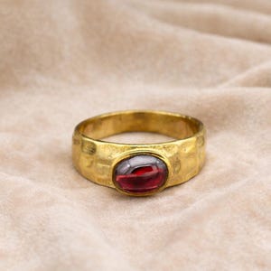 Puede incluir: Un anillo dorado con textura martillada y una gema ovalada de color rojo intenso en el centro. El anillo se encuentra sobre un fondo neutro y texturizado, resaltando su estilo vintage.