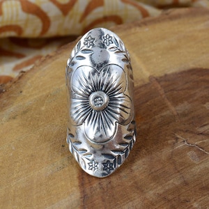 Op de afbeelding: Een zilverkleurige ring met een bloemmotief. De ring heeft een grote bloem in het midden, omringd door kleinere bloemen en bladeren. De ring is gemaakt van een glanzend metaal en heeft een gedetailleerd, getextureerd oppervlak.