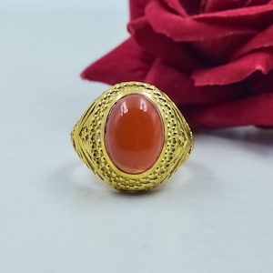 Puede incluir: Un anillo dorado con una piedra ovalada de color rojo anaranjado. El anillo tiene una banda texturizada con un patrón detallado. Una rosa roja está en el fondo. Es una pieza de joyería.