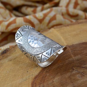 Puede incluir: Un anillo plateado con una superficie texturizada y martillada y patrones geométricos. El anillo presenta una banda ancha con diseños triangulares y punteados, creando un accesorio llamativo. El anillo mide aproximadamente 3,8 cm de largo.