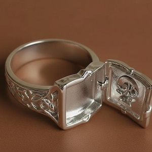 Puede incluir: Un anillo plateado con un diseño floral ornamentado en la banda. El anillo presenta un compartimento rectangular que se abre para revelar un diseño de calavera. El anillo se muestra sobre un fondo marrón.