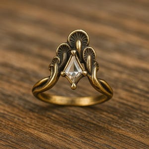 Op de afbeelding: Een ring in vintage-stijl met een heldere, ruitvormige edelsteen gezet in een bronskleurig metaal. De ring heeft sierlijke, paddenstoelachtige details rond de edelsteen en een gladde, gebogen band.