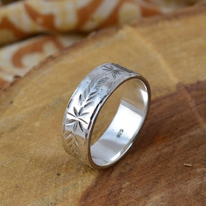 Peut inclure: Une bague en argent avec des motifs gravés de feuilles et d'éclats d'étoiles. La bague est en métal poli et présente une bande avec des motifs détaillés. La bague est exposée sur une surface en bois.