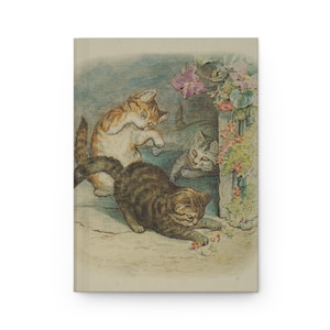 May include: A hardcover journal featuring a vintage illustration of three kittens playing near a stone wall with flowers. The kittens are in shades of brown, orange, and gray, with a soft, watercolor-like style.