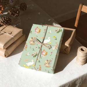 May include: A gift wrapped in light green paper with a whimsical pattern of ducks, flowers, and figures, tied with twine. Other gifts wrapped in brown paper are stacked nearby, with a spool of twine and decorative elements in the background.