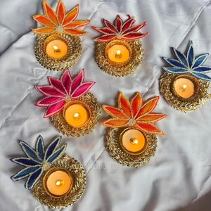 Könnte beinhalten: Eine Sammlung dekorativer Kerzenhalter mit floralem Design. Jeder Halter hat eine goldfarbene Basis mit einer brennenden Kerze und einer farbenfrohen, gestickten Blume in Orange-, Rot-, Rosa- und Blautönen. Die Halter sind auf einer weißen Oberfläche angeordnet.