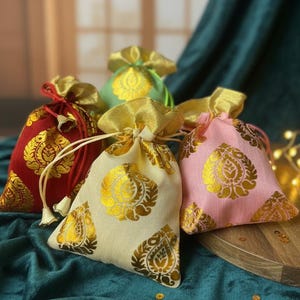 May include: A selection of small drawstring bags in red, green, pink, and cream. Each bag is adorned with a gold paisley design and a gold drawstring. The bags are displayed on a wooden surface against a teal backdrop.