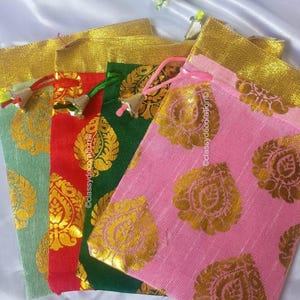 Könnte beinhalten: Eine Auswahl an farbigen Stoffbeuteln mit goldenen Akzenten und Kordelverschlüssen. Die Beutel sind in Grün-, Rot-, Grün- und Rosatönen gehalten und mit einem goldenen Blumenmuster verziert. Im Hintergrund sind weiße Blumen zu sehen.
