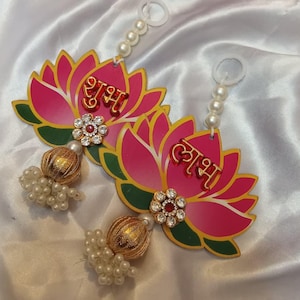 Può includere: Due ornamenti decorativi a forma di fiori di loto con petali rosa, bordi verdi e scritte dorate. Ogni ornamento ha un nappa dorata di perline e un filo di perle. I fiori sono adornati con dettagli di strass e sono su uno sfondo di raso bianco.