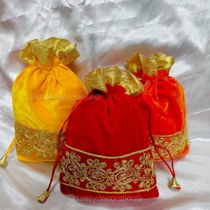 Puede incluir: Tres bolsas de terciopelo con cordón en amarillo, rojo y naranja, con ribetes y cordones bordados en dorado. Las bolsas tienen tela dorada en la parte superior. El texto "@classydecoration" es visible en las bolsas. Almendras esparcidas.