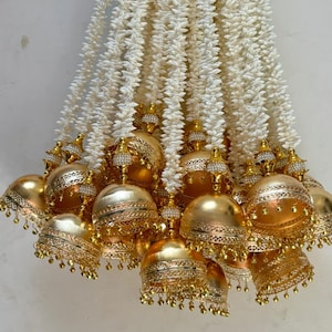 May include: A collection of gold-colored bells with intricate detailing and dangling accents, interspersed with strands of white, textured floral elements. The bells have a shiny finish and are arranged in a decorative display.