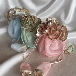 May include: Three pastel-coloured drawstring pouches: light blue, mint green, and pink. Each pouch has a ruffled, sequined gold trim. The drawstring matches the pouch colour, with a small, sequined pouch attached.