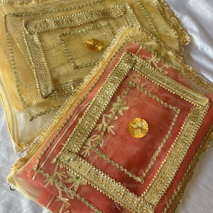 May include: Two decorative, square-shaped textiles. One is a light peach color, and the other is a golden yellow. Both have intricate gold trim and a central, circular gold embellishment. The textiles are likely used for decorative purposes.