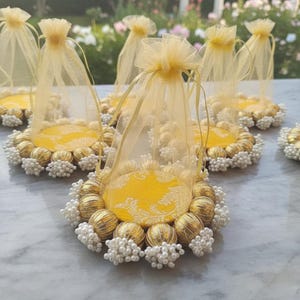 May include: Several decorative favor bags with yellow fabric bases, gold-colored spheres, and white pearl accents. Each bag has a sheer, light yellow drawstring pouch. The items are arranged on a light-colored surface, with a blurred outdoor background.