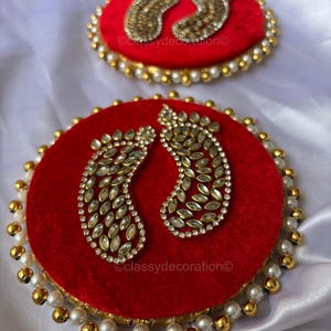 Può includere: Due pezzi decorativi in velluto rosso con finiture in oro e perle. Ogni pezzo presenta un paio di impronte decorate con accenti chiari e dorati. Il testo "@classydecoration" è visibile.