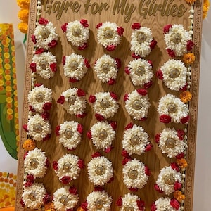 May include: A wooden board with gold lettering reading "Gajre For My Girlies". It features multiple white flower arrangements with red and gold accents, and orange flowers, arranged on the board.