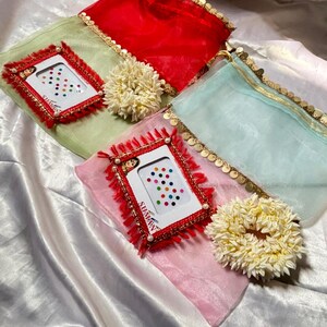 May include: An assortment of colorful items, including red, green, pink, and blue sheer scarves with gold trim. Each scarf is paired with a small, framed card with the text "SUMAN" and a variety of colorful dots, along with a cream-colored floral hair accessory.