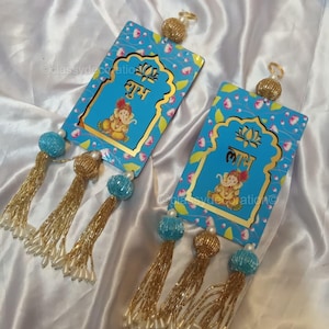Könnte beinhalten: Zwei rechteckige blaue Dekohänger mit goldenen Rändern, jeweils mit einer Ganesha-Illustration und Hindi-Text. Sie haben goldene Perlenaufsätze, türkisfarbene Perlen und goldene Quasten mit weißen Perlen. Floralmuster im Hintergrund.