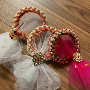 Puede incluir: Tres diademas decorativas con un diseño circular. Cada diadema presenta una tela de red y está adornada con perlas, detalles dorados y joyas coloridas. Una tiene una red blanca, otra una red rosa y la tercera una red blanca.