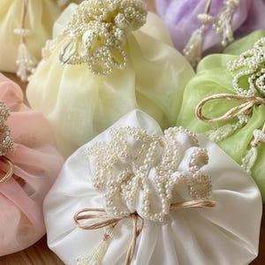 May include: A collection of colorful, fabric pouches in shades of white, yellow, pink, green, and purple. Each pouch is adorned with a cluster of faux pearls and tied with a natural twine bow, creating an elegant and delicate aesthetic.